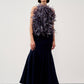 A model stands in a studio wearing a sleeveless dark navy maxi dress with a voluminous dark purple feathered bodice detail, paired with nude high heels.