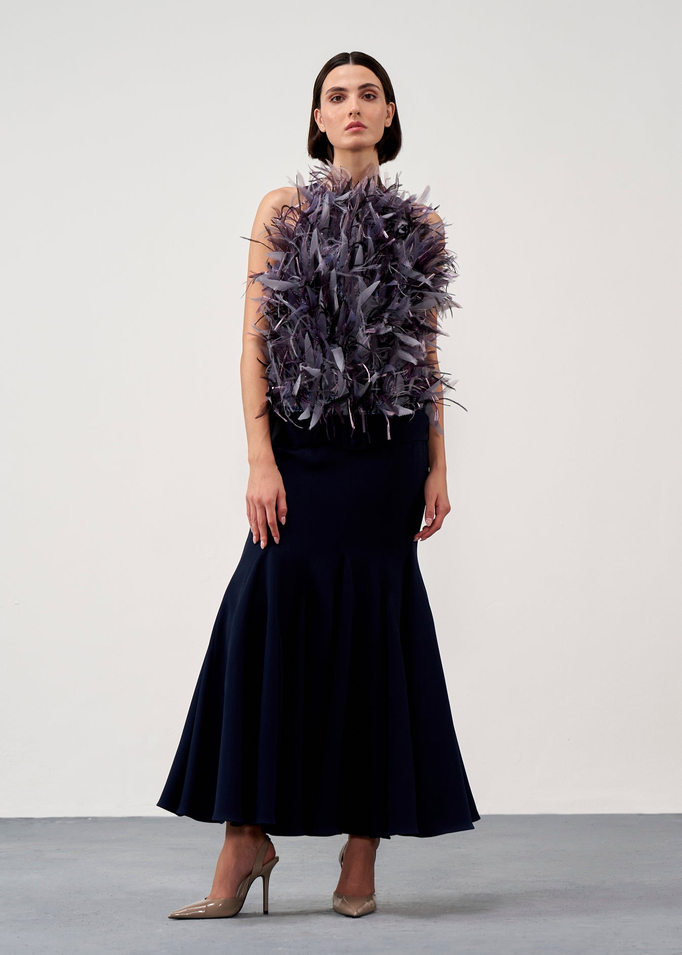 A model stands in a studio wearing a sleeveless dark navy maxi dress with a voluminous dark purple feathered bodice detail, paired with nude high heels.