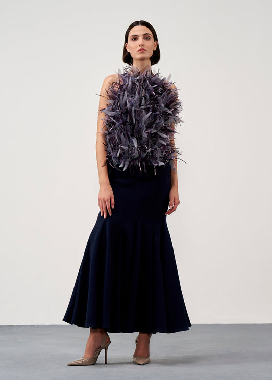 A model stands in a studio wearing a sleeveless dark navy maxi dress with a voluminous dark purple feathered bodice detail, paired with nude high heels.